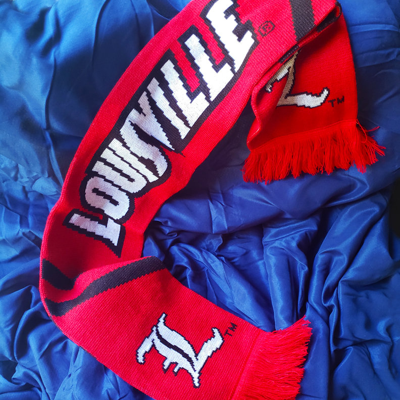 A scarf with University of Louisville's old english L and the word Louisville knitted along the length of it.